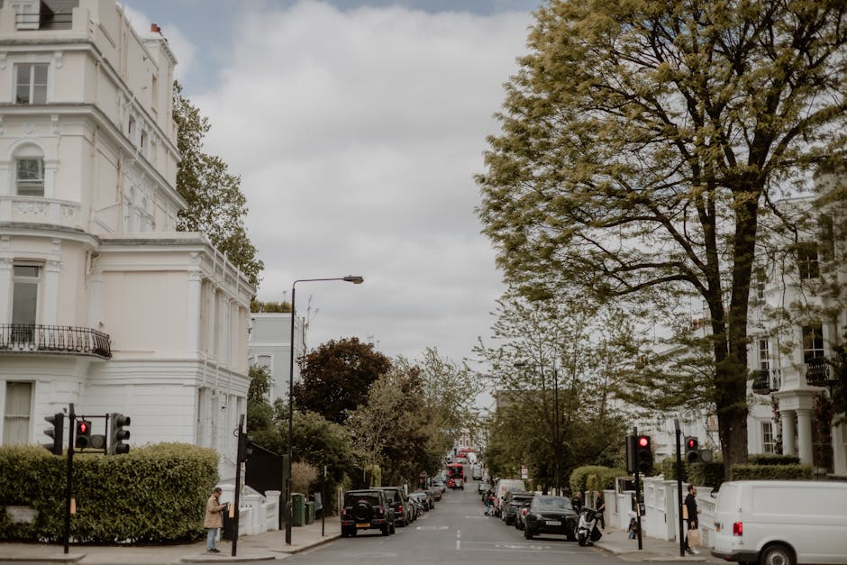 A wide view of a residential street in Belsize Park, showing white Victorian-style buildings with detailed facades and balconies on each side. Several parked cars, including a black van, are lined up 