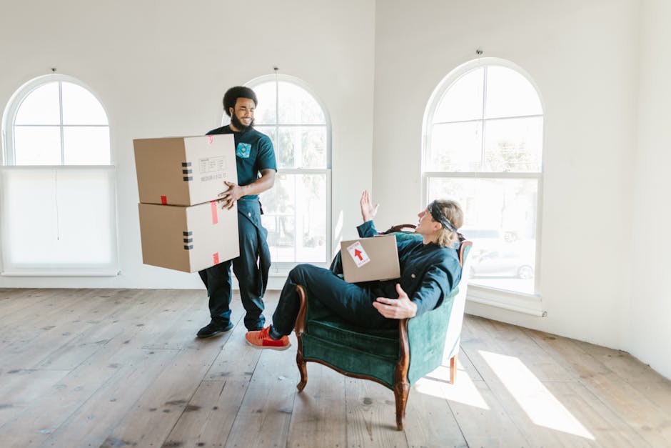 A man with curly hair and a beard, dressed in a blue t-shirt and dark pants, is seated on a light wooden floor inside a room with two large arched windows that let in natural daylight. He is surrounde